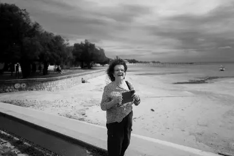 Elizabeth, 2022. A woman laughing on a beach promenade, the tide out behind her, in black and white.